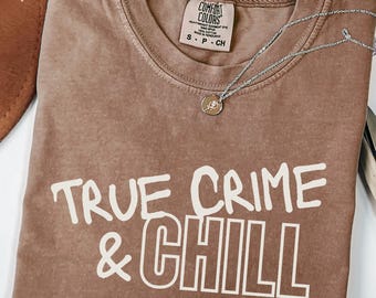 Op de afbeelding: Espresso-kleurig T-shirt met de tekst "TRUE CRIME & CHILL" in witte blokletters. Het shirt heeft een ronde hals en een kleine zilveren ketting met een hanger. Het shirt is gemaakt van Comfort Colors materiaal.