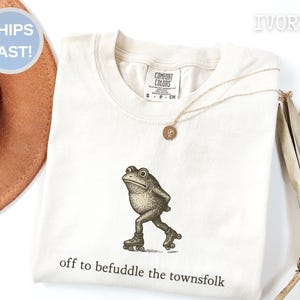 Könnte beinhalten: Elfenbeinfarbenes T-Shirt mit einer Vintage-Illustration eines Frosches auf Rollschuhen. Unter dem Frosch steht der Text "off to befuddle the townsfolk". Eine goldene Halskette mit Anhänger liegt auf dem Shirt.