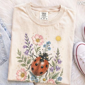 Può includere: Una t-shirt color avorio con una stampa grafica di una grande coccinella rossa con punti neri circondata da fiori rosa e viola, foglie verdi e un sole giallo. La maglietta presenta un girocollo e un'etichetta del marchio Comfort Colors.