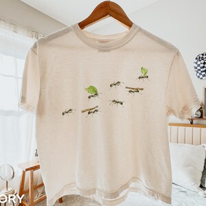 Könnte beinhalten: Ein elfenbeinfarbenes Kurzarm-T-Shirt mit einem Grafikdruck von mehreren Ameisen, die kleine grüne Blätter und braune Zweige über die Brust tragen. Das T-Shirt hat einen Rundhalsausschnitt und wird auf einem Holzkleiderbügel vor hellem Hintergrund präsentiert.