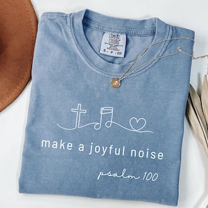 May include: A blue Comfort Colors t-shirt with the text "make a joyful noise psalm 100" and a cross, musical note, and heart design. A gold necklace and a brown hat are also visible.