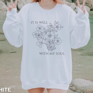 Puede incluir: Sudadera blanca con el texto "IT IS WELL WITH MY SOUL" y un diseño floral y de mariposa. La sudadera tiene cuello redondo y mangas largas. La palabra "WHITE" está impresa en la parte inferior de la imagen.