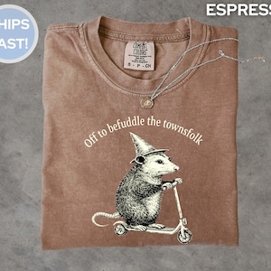 Könnte beinhalten: Espressofarbenes T-Shirt mit einer Grafik eines Opossums auf einem Roller und einem Zaubererhut. Der Text "Off to befuddle the townsfolk" ist über dem Opossum angeordnet. Eine silberne Halskette mit Anhänger liegt auf dem Shirt.
