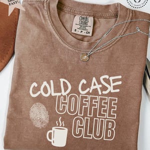 May include: Espresso-colored t-shirt with the text "COLD CASE COFFEE CLUB" in white. The design includes a fingerprint and a coffee mug graphic. A silver necklace with a pendant is draped across the shirt.