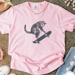 Puede incluir: Camiseta rosa claro con un gr&aacute;fico de un gato atigrado gris montando en monopat&iacute;n negro. El gato es el foco principal. La camiseta tiene cuello redondo y mangas cortas. Un collar dorado rodea el cuello.