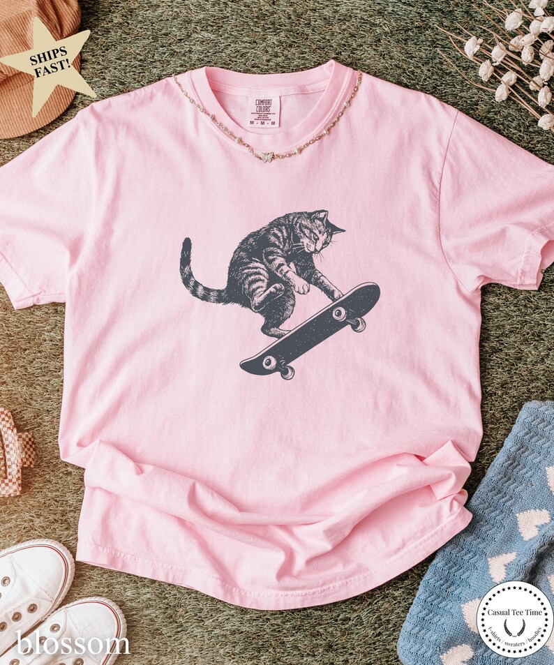 Puede incluir: Camiseta rosa claro con un gr&aacute;fico de un gato atigrado gris montando en monopat&iacute;n negro. El gato es el foco principal. La camiseta tiene cuello redondo y mangas cortas. Un collar dorado rodea el cuello.