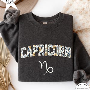 May include: Dark heather gray sweatshirt with the word "CAPRICORN" in a floral design across the chest. Below the word is the Capricorn zodiac symbol in white. The text "dark heather" is at the bottom.