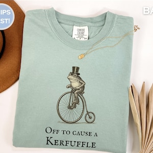 Peut inclure: T-shirt bleu clair avec une illustration fantaisiste d'une grenouille en chapeau haut de forme sur un grand-bi. Le texte "Off to cause a kerfuffle" est imprimé en dessous. Un collier doré est posé sur le t-shirt.