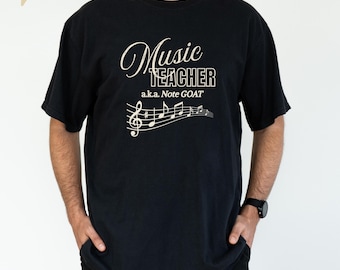 May include: Black t-shirt with the text "Music Teacher a.k.a. Note GOAT" in a cream-colored script font. A musical staff with notes is below the text. The shirt is a casual style.