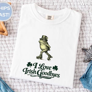 Könnte beinhalten: Weißes T-Shirt mit einer grünen Froschfigur, die einen Zylinder und Rollschuhe trägt. Der Text "I Love Irish Goodbyes" ist darunter in Grün gedruckt. Das T-Shirt ist auf einer weißen Oberfläche gefaltet.