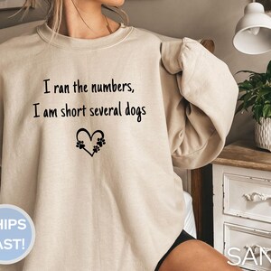 May include: A sand-coloured sweatshirt featuring the text "I ran the numbers, I am short several dogs" in black. A heart shape with paw prints is below the text. The sweatshirt has a relaxed fit.