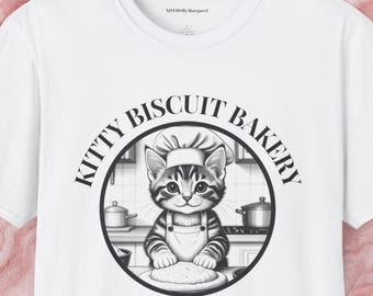 May include: White t-shirt featuring a circular graphic of a cat wearing a chef's hat and apron, with the text "KITTY BISCUIT BAKERY" around the cat. The text "ESTABLISHED IN 1988" is below the graphic. The brand name "NDTHS Margaret" is at the top.