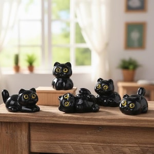 May include: A collection of five glossy black cat figurines with yellow eyes. The cats are in various poses, including sitting, lying down, and standing. They are arranged on a wooden surface, creating a charming display.