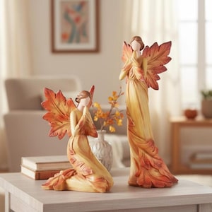 May include: Two angel figurines with leaf-shaped wings. One kneels, the other stands. Both wear long, flowing gowns in shades of yellow and orange, with matching leaf-shaped wings. The figurines are on a light grey table, with a neutral background.