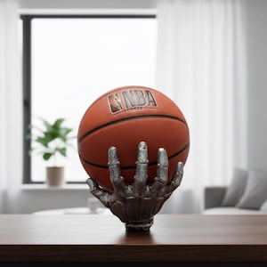 May include: An orange basketball with the NBA FORGE logo is held by a silver skeleton hand sculpture. The sculpture is on a wooden table. The basketball has black lines.