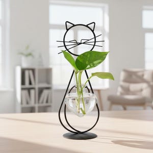 May include: A black metal cat-shaped plant holder with a clear, heart-shaped glass vase. The vase holds green leafy plants. The cat's face is a simple wireframe design, with a round head, ears, and whiskers. The holder sits on a round base.