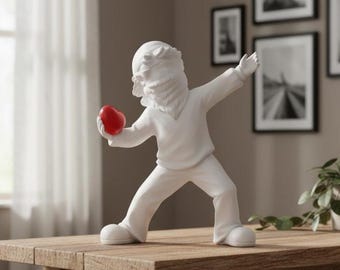 May include: A white ceramic sculpture of a figure throwing a red heart. The figure is wearing a white sweater, pants, and shoes. The sculpture is on a wooden table. Decorative art for home decor.