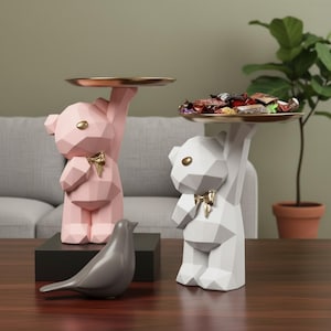 May include: Two geometric bear ornaments, one pink and one white, each holding a gold-coloured tray. The pink bear has a gold bow, and the white bear holds a tray of wrapped sweets. A grey bird ornament sits on a black base.