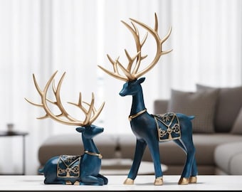 May include: Two decorative deer figurines. One is standing, the other is lying down. Both are dark blue with gold-colored antlers and accents. The standing deer has a decorative saddle. The figurines are on a white surface.