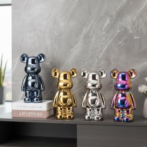 May include: Four metallic bear figurines in blue, gold, silver, and iridescent colours. Each bear has a heart-shaped detail on its chest. The figurines are displayed on a grey surface, with a book that reads "SUNLIGHT" in white.