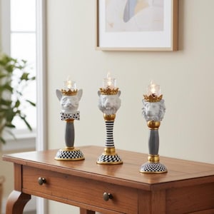 May include: Three decorative candle holders with animal head designs. Each holder features a different animal head, a gold crown, and a small glass candle holder. The bases have black and white checkered patterns, with gold and gray accents.