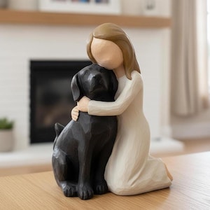 May include: A figurine of a person in a white dress kneeling and hugging a black dog. The person has light brown hair and a faceless design. The dog is a Labrador Retriever. The figurine is on a wooden table.