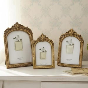 May include: Three gold-coloured picture frames with elaborate detailing. Each frame has an arched top and a floral design. The frames have a white background with a small square opening for a photo. The frames are on a white surface.