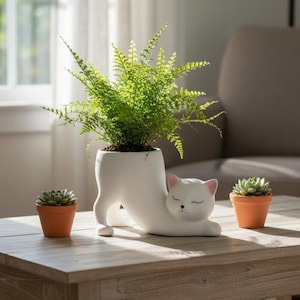 May include: A white ceramic cat-shaped planter holding a green fern. Two small terracotta pots with succulents sit nearby. The cat planter has pink accents on the ears and closed eyes. The scene is set on a wooden table.