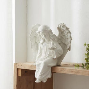 May include: A white angel figurine with detailed wings and flowing robes. The angel is seated, holding a small bird in its hands. The sculpture is placed on a wooden shelf, creating a serene and decorative display.