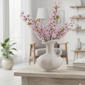 May include: A white ceramic vase with a handle holds a bouquet of pink cherry blossom branches. The vase sits on a light-colored wooden table, with a chair and other decor in the background.