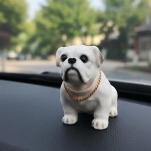 May include: A white bulldog figurine with a gold-colored chain necklace. The dog has black eyes and a black nose. The figurine is sitting on a black surface, with a blurred background of trees and a building.