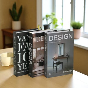 May include: Three decorative books with hard covers. The book spines display the words "DESIGN" and "DECOR". The first book has the text "VANITY FAIR 100 YEARS" and "FROM THE JAZZ AGE TO". The third book has the text "Boconcept Collection Must".