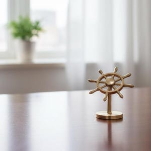 May include: A gold-colored ship's wheel decorative object. The wheel has eight spokes and is mounted on a small, round base. The object is displayed on a dark wooden surface, with a blurred background of a window and a potted plant.