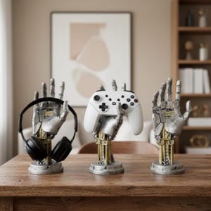 May include: Three robotic hand-shaped display stands. One holds black headphones, another a white game controller, and the third is empty. The stands are silver and gold, set on a wooden table. The background includes a framed artwork and a bookshelf.