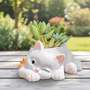 May include: A white cat-shaped planter with a winking eye, holding a small orange flower. The planter is filled with green succulents. The cat is lying down on a wooden surface, with a blurred garden background.