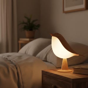 May include: A wooden bird-shaped table lamp emitting a warm glow. The lamp features a light-coloured, rounded body and darker wooden head and wings. It rests on a wooden stand, positioned on a wooden bedside table next to a bed.