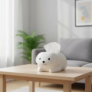 May include: A white, dog-shaped tissue box dispenser with black eyes and a small black nose. White tissues emerge from the top. It sits on a light brown wooden table. The background includes a grey sofa and a green plant. The tissue box is made of ceramic.