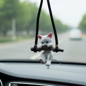 May include: A white cat figurine with pink ears and blue eyes hangs from a black cord. The cat is perched on a brown branch, designed as a car accessory. The background shows a blurred road and a car dashboard.