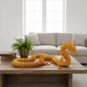 May include: An orange dragon sculpture with intricate details rests on a wooden coffee table. The dragon is posed with its head raised and body coiled. A potted fern and books are also on the table, with a neutral-toned sofa and window in the background.