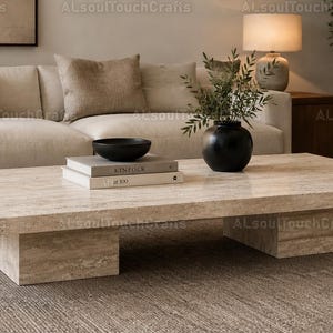 May include: A rectangular, light-coloured stone coffee table with a minimalist design. The table has a smooth surface and is supported by two rectangular bases. A black bowl, books, and a black vase with greenery are on the table.