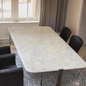 May include: A rectangular dining table with a white, marble-like surface, supported by a dark base. Six black chairs surround the table, set on a patterned rug. The room has a window and curtains.