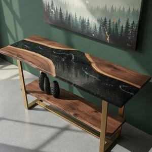 Puede incluir: Una consola de madera hecha a mano con un diseño de río de resina epoxi negra. La mesa tiene un estante inferior y patas de metal de color dorado. Tres jarrones negros decorativos están en el estante. Una pintura de un bosque cuelga en el fondo.
