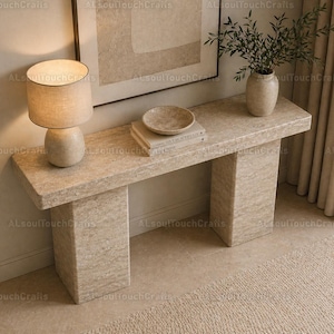 Puede incluir: Una consola de piedra beige con dos patas cuadradas. Una lámpara beige con una pantalla cilíndrica se encuentra sobre la mesa, junto con un cuenco pequeño, una pila de libros y un jarrón con vegetación. Una gran obra de arte enmarcada cuelga de la pared.