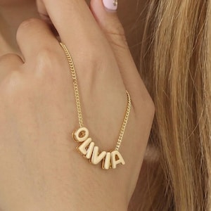 May include: A gold-colored necklace with the name "OLIVIA" spelled out in individual letters. The necklace is on a delicate chain and is being held up by a hand with a pink nail polish.