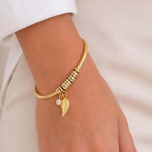 Puede incluir: Una pulsera dorada con un dije de hoja y una pequeña gema transparente. La pulsera tiene una serie de pequeñas cuentas oscuras y se usa en la muñeca. El fondo muestra a una persona con una camisa blanca y pantalones color crema.