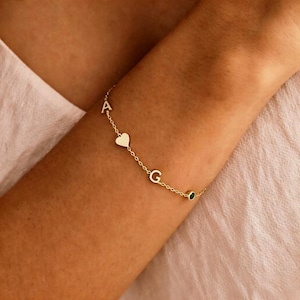 May include: Gold-colored bracelet with the letters "A" and "G", a heart charm, and a small green gemstone. The bracelet is delicate and appears to be made of linked chain. The bracelet is worn on the arm.