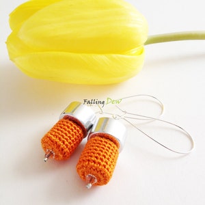Puede incluir: Un par de pendientes de crochet naranja con detalles en metal plateado. Los pendientes cuelgan de ganchos plateados. La palabra "Falling Dew" es visible en el fondo.