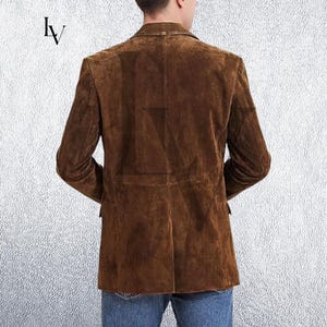 May include: A brown suede blazer with a classic two-button closure. The jacket features a notched lapel and two front pockets. The back view shows the tailored fit and the soft texture of the suede. The blazer is worn with blue jeans.
