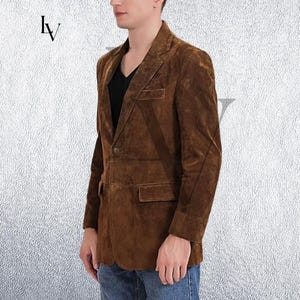 May include: A brown suede blazer with a notched lapel, two front pockets, and a single-button closure. The jacket is worn over a black shirt and blue jeans. The blazer has a classic, tailored fit.