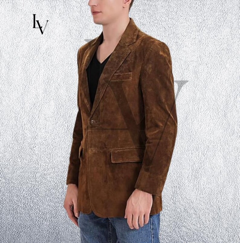 May include: A brown suede blazer with a notched lapel, two front pockets, and a single-button closure. The jacket is worn over a black shirt and blue jeans. The blazer has a classic, tailored fit.